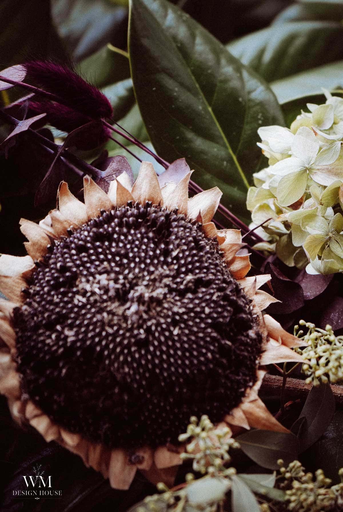 Close-up of a sunflower with dark seeds, surrounded by lush green leaves and other blooms, including a pale hydrangea, evoking the fresh elegance of a greenery table runner. The WM Design House logo is visible in the lower left corner.