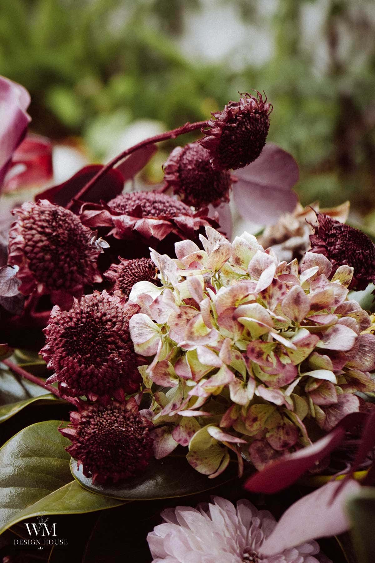 A close-up of a floral arrangement featuring pale pink hydrangeas and dark burgundy flowers with leafy greens, beautifully reminiscent of a lush greenery table runner. The WM Design House logo appears in the bottom left corner.