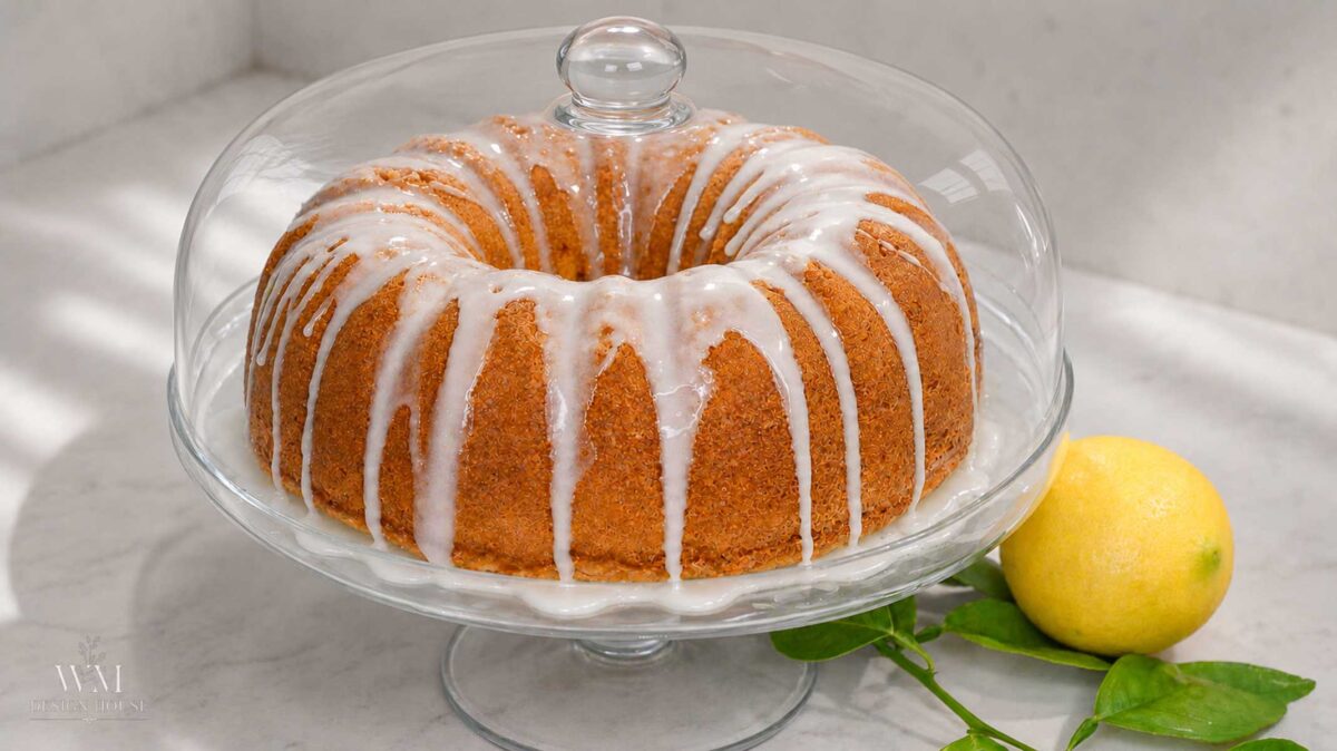 A lemon bundt cake with white glaze drips sits on a glass cake stand with a dome lid. A whole lemon with leaves is placed next to the stand on a light countertop.