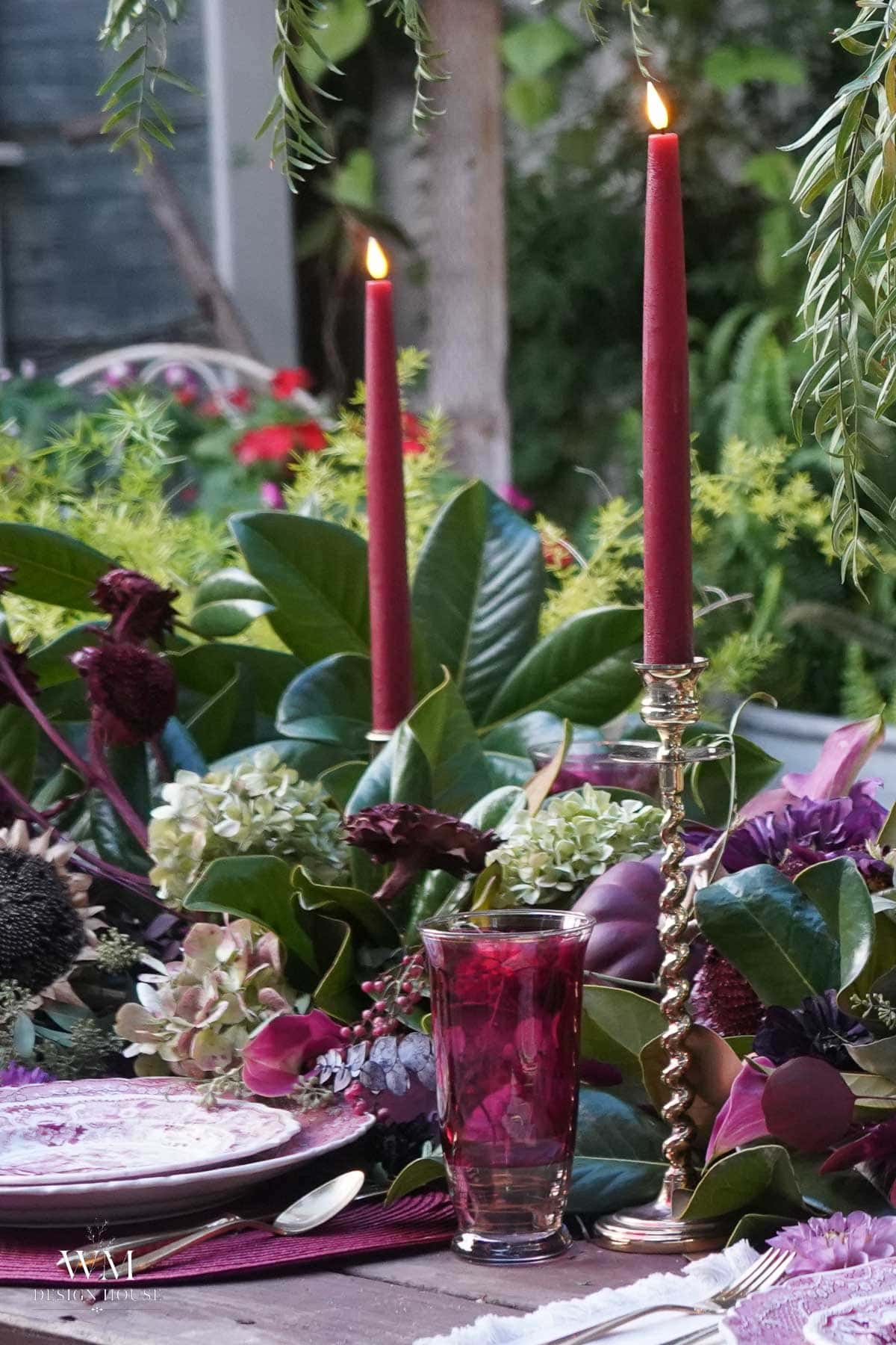 A close-up of a table set outdoors with two tall red candles, a red glass, floral arrangements, and elegant plates on display, featuring a lush greenery table runner and surrounded by vibrant flowers.