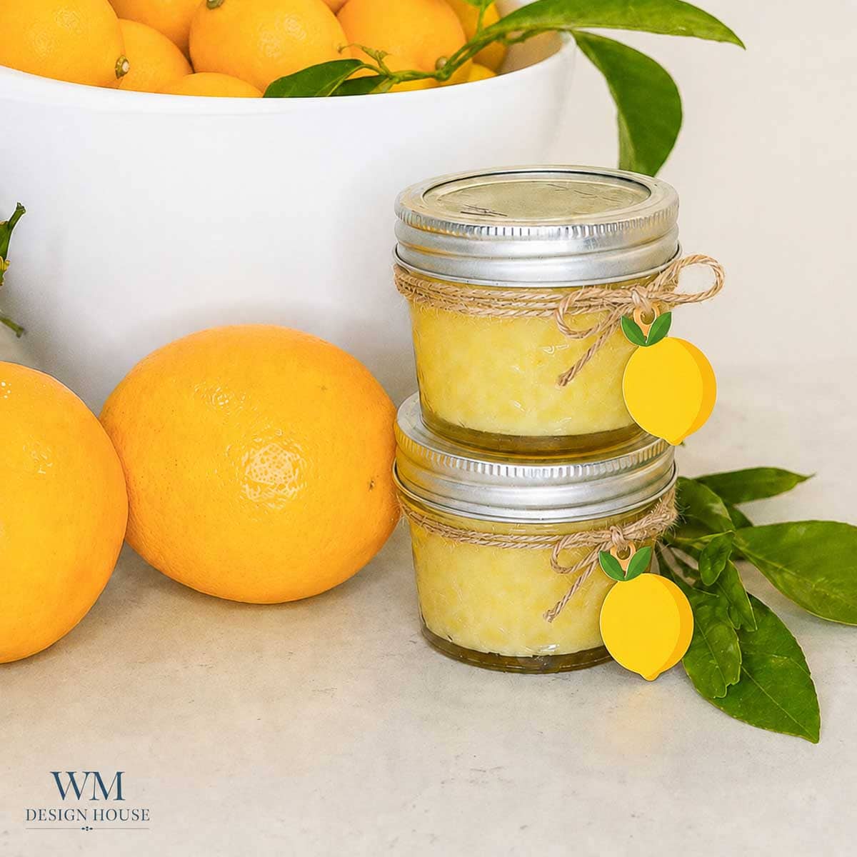 Two small jars of Meyer Lemon Curd are stacked, tied with twine and decorated with lemon-shaped tags. Fresh lemons and a bowl filled with lemons and green leaves sit beside them on a white surface. WM Design House logo appears in the corner.
