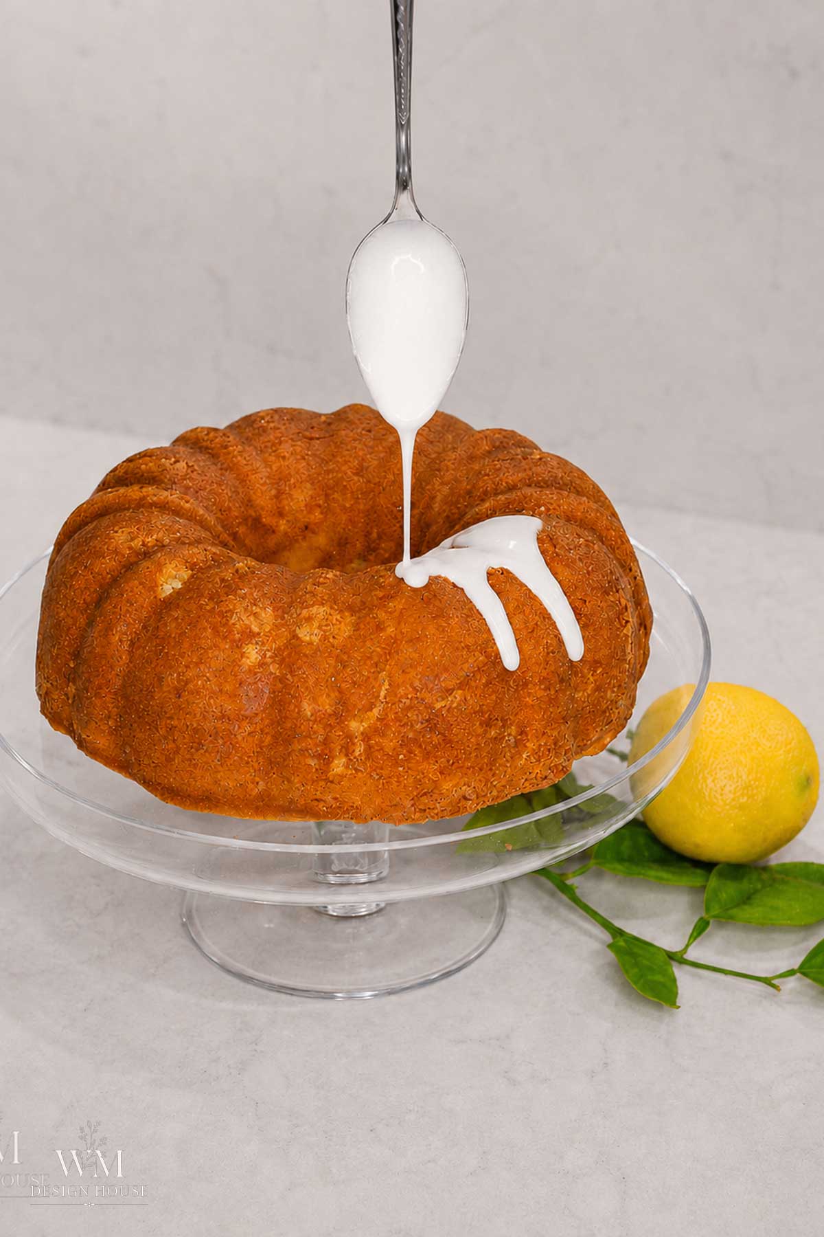 A spoon drizzles white icing onto a bundt cake on a glass stand. A lemon with leaves sits nearby on a light-colored surface.