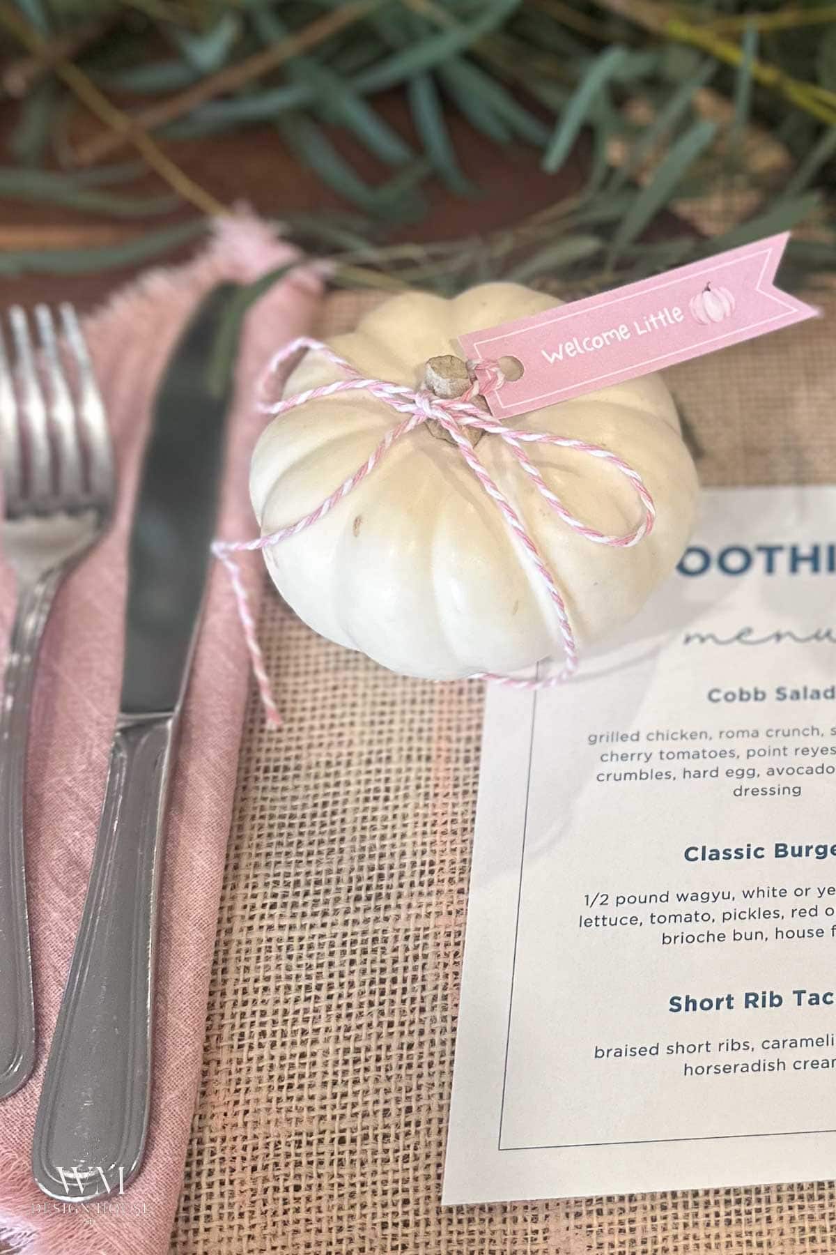 A small white pumpkin with a “Welcome Little” pink tag sits next to a fork and knife on a pink napkin, with part of a printed menu visible on a burlap tablecloth.