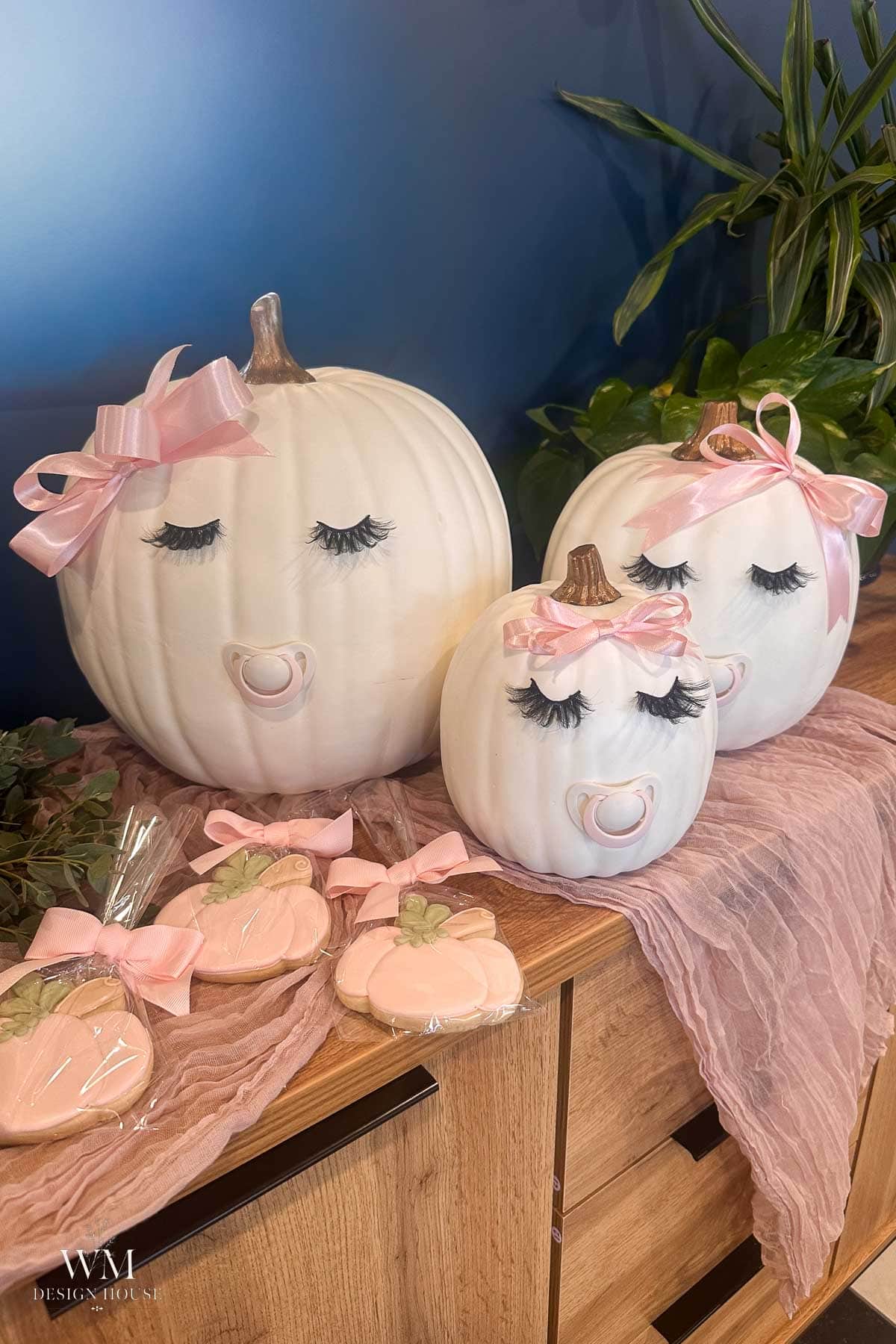 Three white pumpkins decorated as baby faces with pacifiers, pink bows, and eyelashes sit on a pink cloth, next to pumpkin-shaped cookies and green plants on a wooden surface.