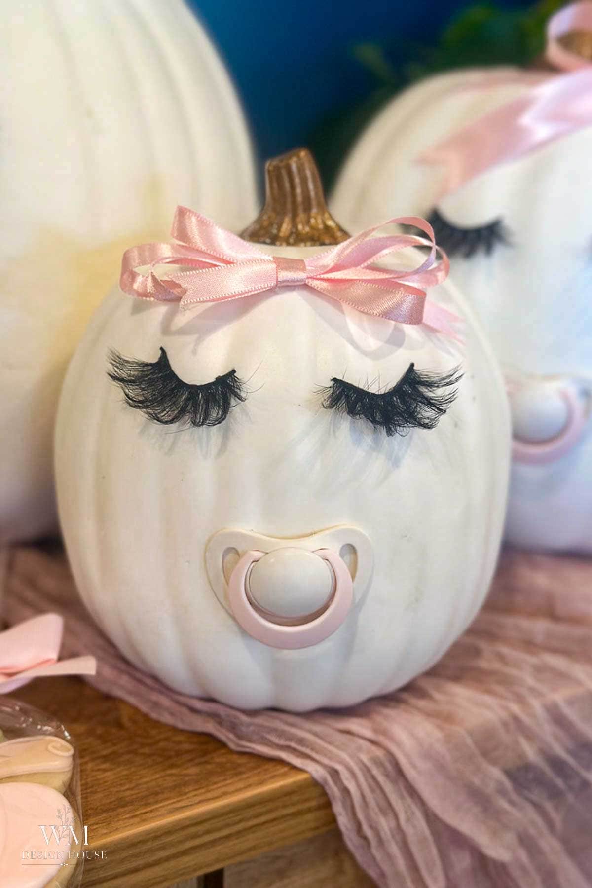 A white pumpkin decorated as a baby with long fake eyelashes, a pink pacifier, and a pink ribbon bow on top, sitting on a pink cloth.