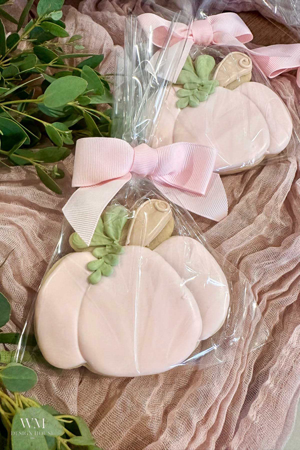 Two pastel pink pumpkin-shaped cookies, decorated with green leaves and wrapped in clear plastic, are tied with a light pink ribbon bow. They rest on a soft pink fabric with green foliage nearby.