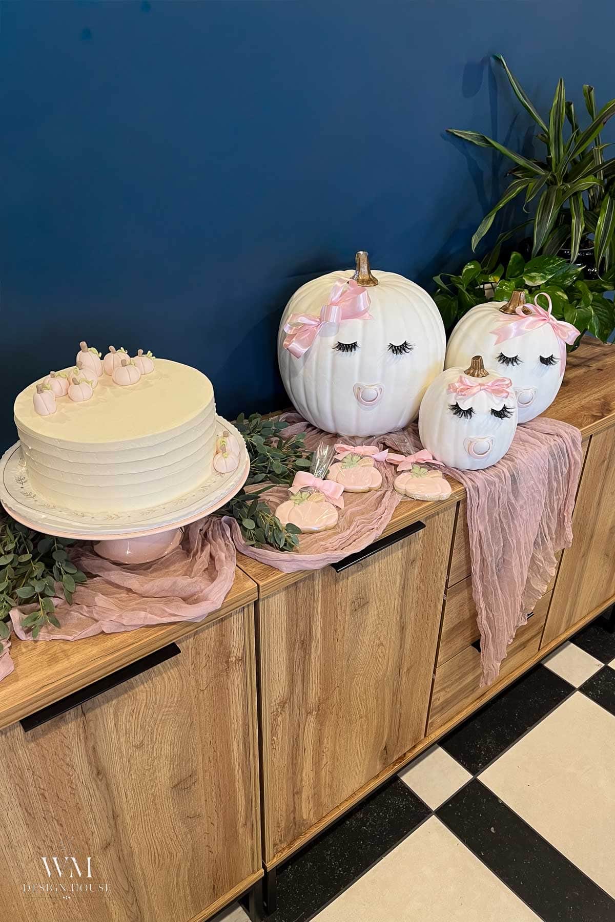 A white cake with pink decorations and three white pumpkins painted with faces and pink bows, displayed on a wooden cabinet with greenery and pink fabric against a blue wall.