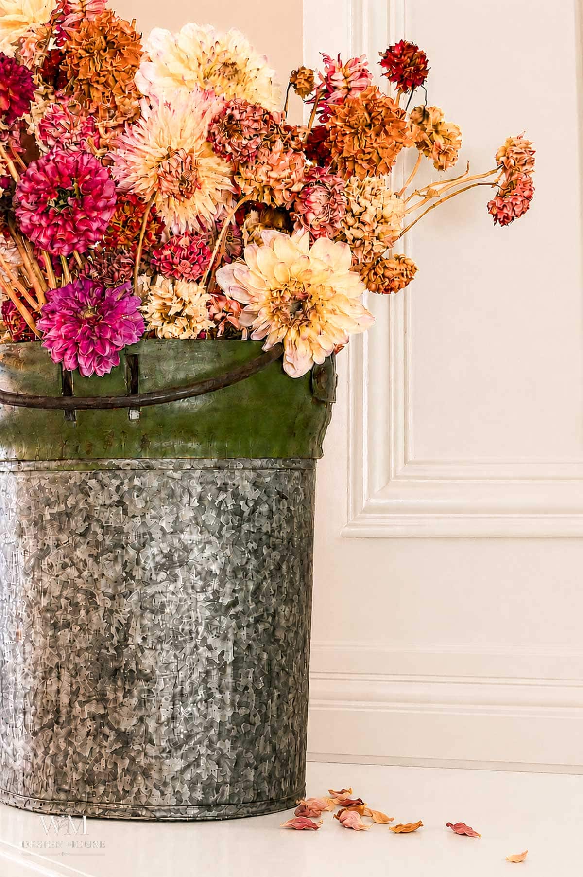 A galvanized metal bucket filled with pink, yellow, and orange dried flowers—perfect for anyone learning how to dry dahlias—sits on a white floor beside an off-white paneled wall. A few dried petals are scattered on the floor.