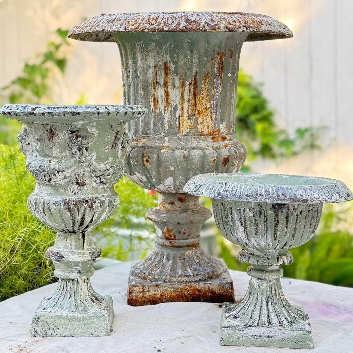 Three ornate, weathered metal urn planters with a green patina and rust—showcasing a beautiful DIY aged urn finish—sit on a stone surface outdoors, surrounded by greenery and a white fence in the background.