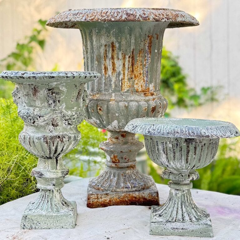 Three ornate, weathered metal urn planters with a green patina and rust—showcasing a beautiful DIY aged urn finish—sit on a stone surface outdoors, surrounded by greenery and a white fence in the background.