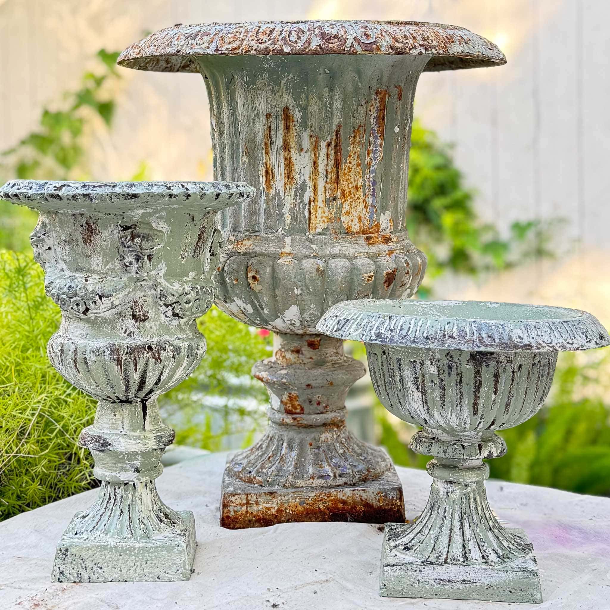 Three ornate, weathered metal urn planters with a green patina and rust—showcasing a beautiful DIY aged urn finish—sit on a stone surface outdoors, surrounded by greenery and a white fence in the background.
