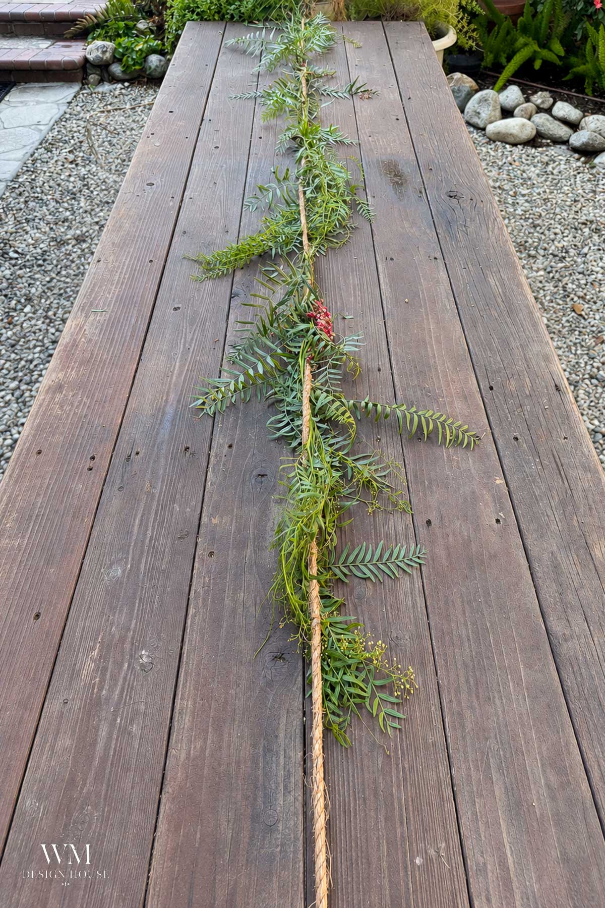 A long garland made of greenery and rope lies on a wooden outdoor table, with rocks and plants in the background.