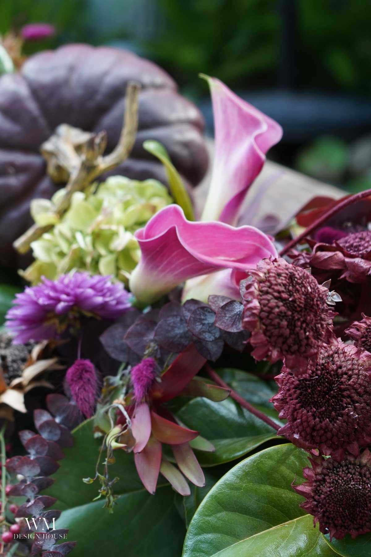 Close-up of a floral arrangement featuring pink calla lilies, purple chrysanthemums, deep red scabiosa, green hydrangeas, dark foliage, and a purple pumpkin in the background.