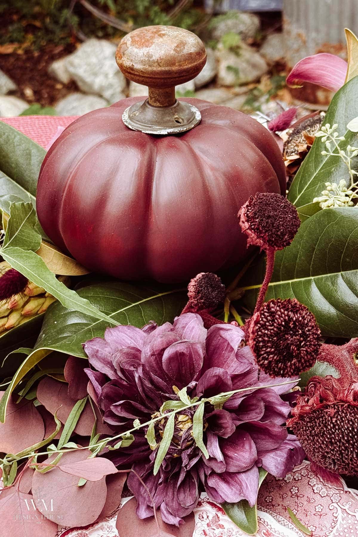 A burgundy pumpkin-shaped object with a metal knob sits among purple flowers, dark red dried blooms, and green leaves atop a lush greenery table runner, creating a rich, autumnal arrangement.