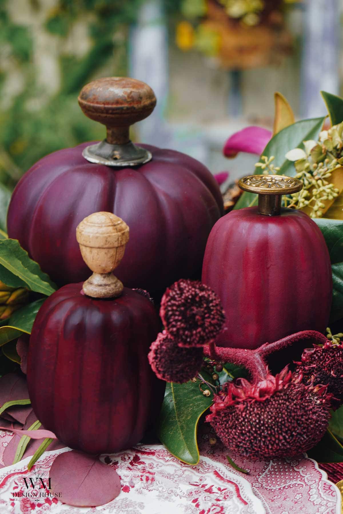 Three decorative bottles shaped like deep purple pumpkins with ornate stoppers, surrounded by green leaves and red textured flowers, arranged on a patterned cloth.
