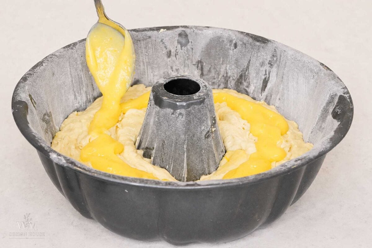 A spoon spreads yellow filling over cake batter in a floured bundt pan, preparing it for baking.