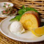 A slice of lemon bundt cake with white glaze, a scoop of vanilla ice cream, and a lemon wedge on a white plate. A floral teacup and woven basket are in the background.