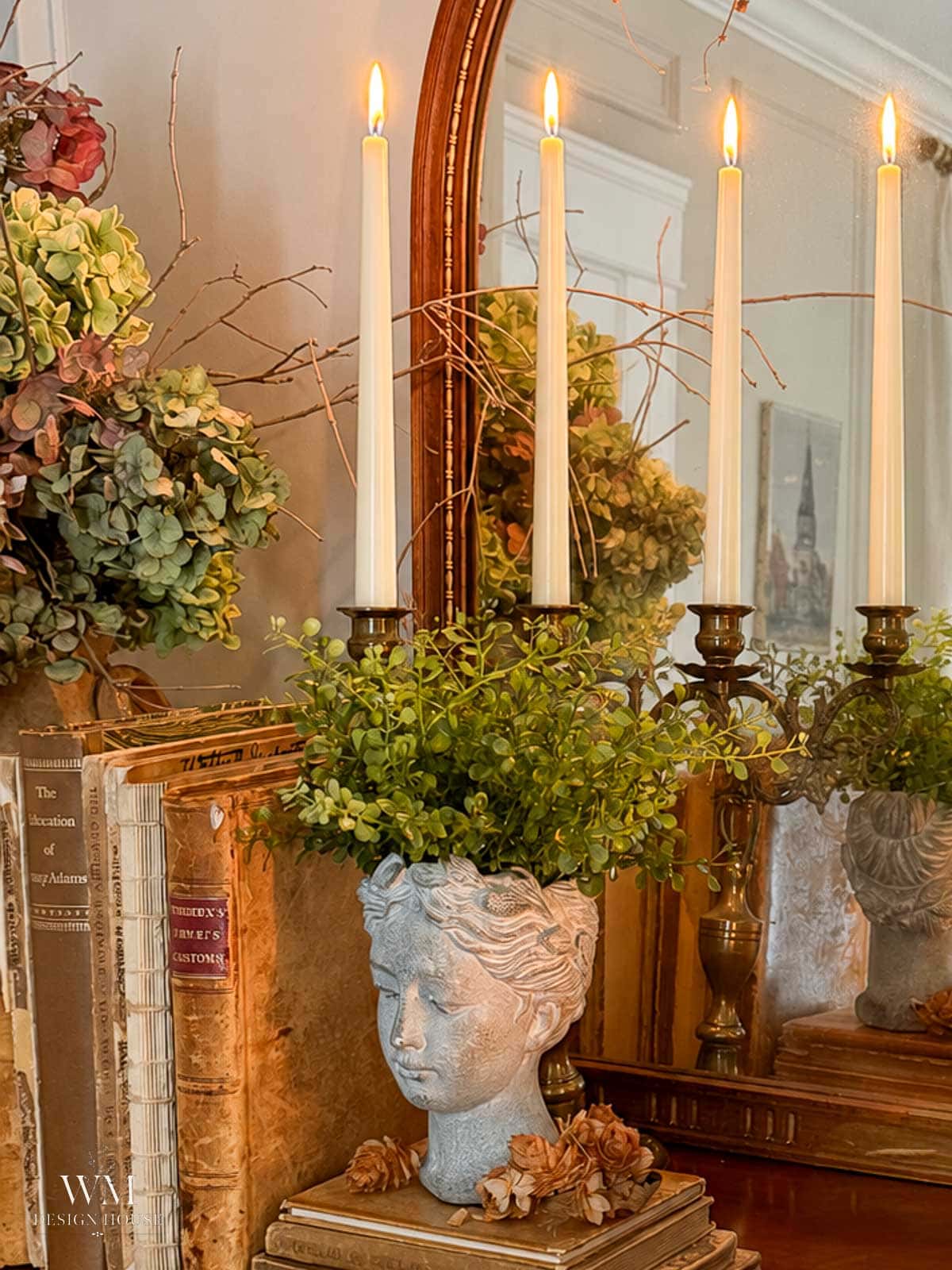 A decorative bust planter with greenery and dried flowers sits on books beside a mirror, adding a touch of fall bedroom decor. Four lit taper candles in a holder are reflected in the mirror, with a bouquet of hydrangeas in the background—perfect for a seasonal refresh or creating a cozy fall bedroom atmosphere.