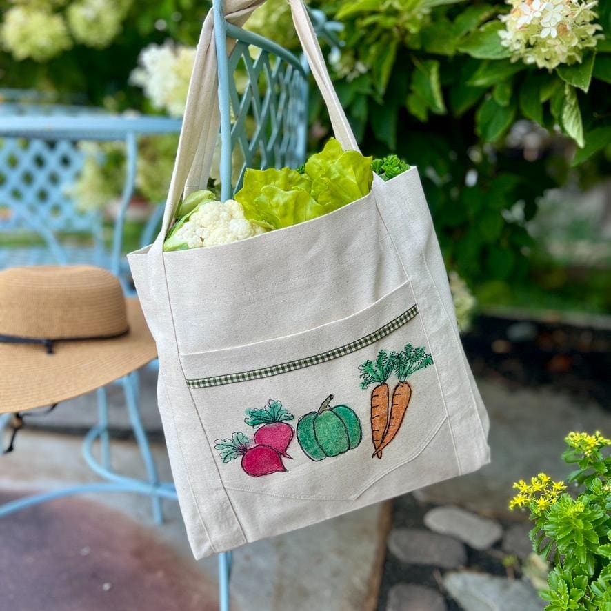 A white tote bag with embroidered vegetables holds fresh greens. The bag hangs on a blue chair outdoors with greenery, flowers, and a straw hat nearby.