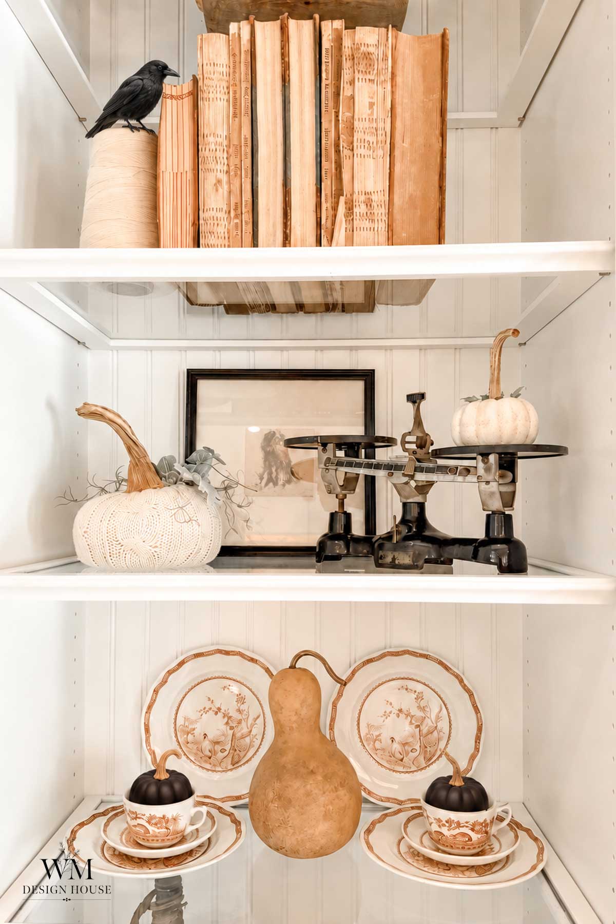Three white shelves display vintage books, a black bird figurine, pumpkins, scales, framed art, and decorative china—including plates, cups, and a gourd. The arrangement is elegant and autumn-themed, with touches like Halloween lanterns DIY projects adding a handcrafted seasonal charm.
