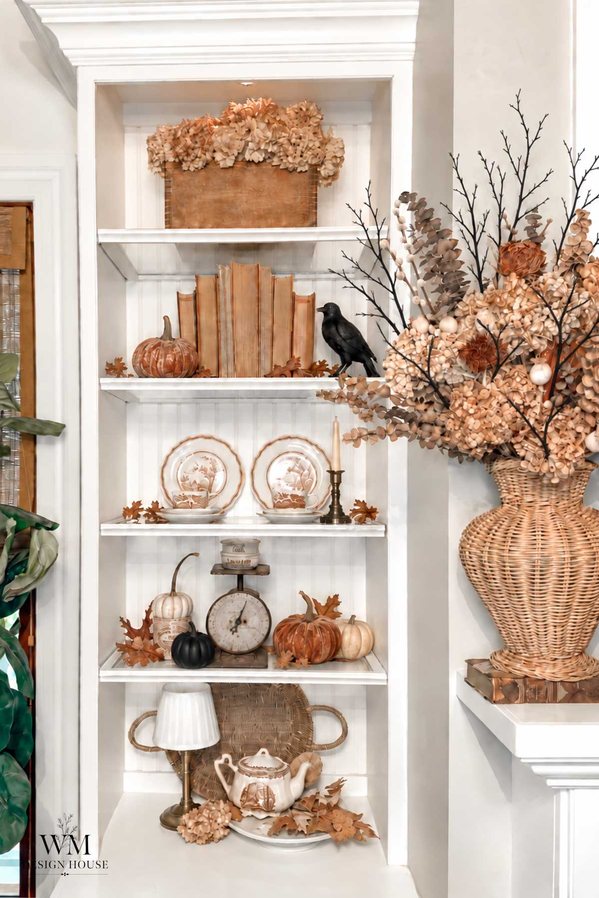 A white corner shelf displays books, pumpkins, vintage dishes, a scale, and a black bird. Dried flowers and leaves add autumn decor, with a large woven vase of hydrangeas on the side. For an extra festive touch, consider adding Halloween lanterns DIY to infuse creative charm into your seasonal display.