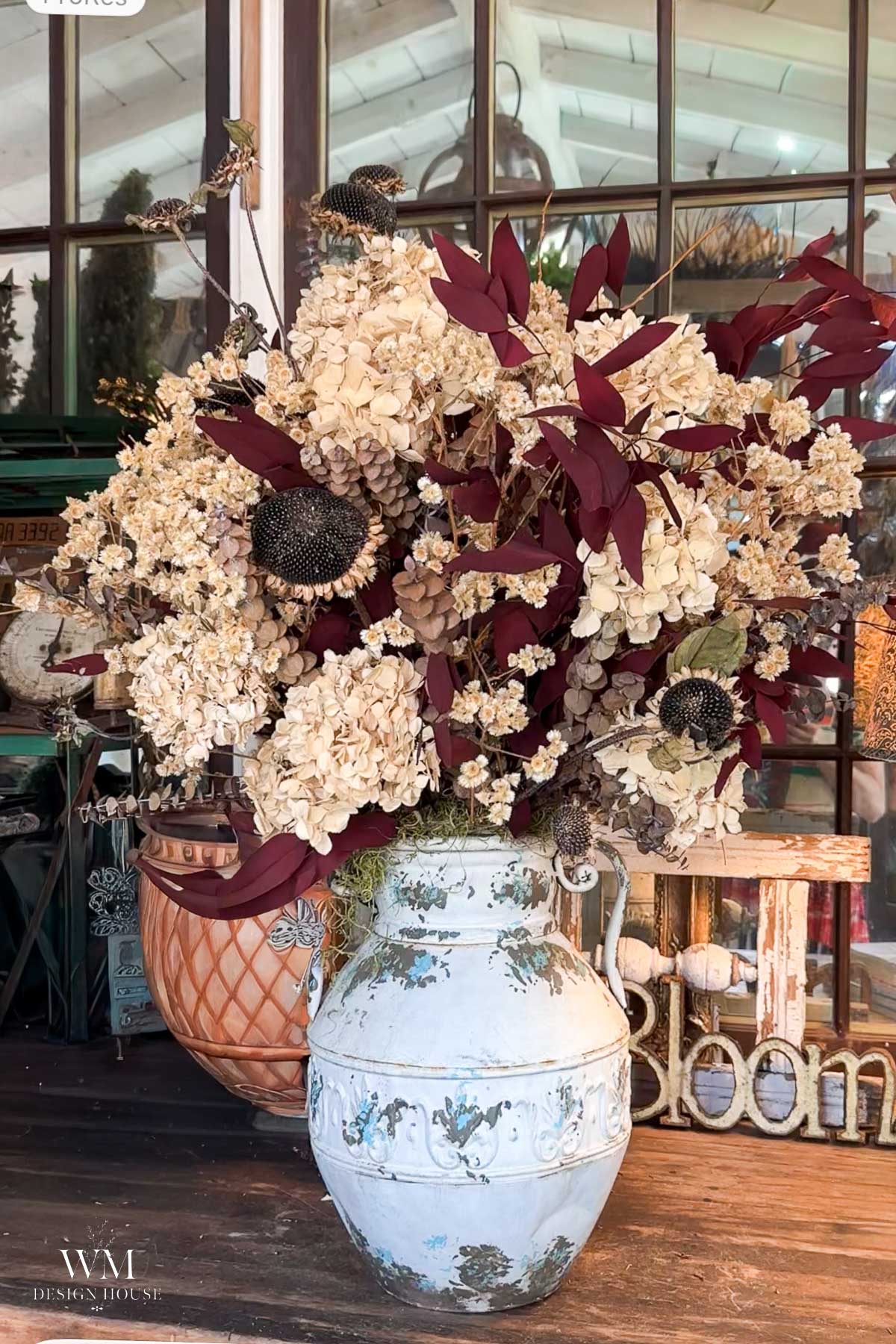 A large ceramic vase holds a dried floral arrangement with cream hydrangeas, dark red leaves, and dried seed pods, placed on a rustic wooden table in a bright, airy room—perfectly complementing festive touches like halloween lanterns DIY scattered nearby.