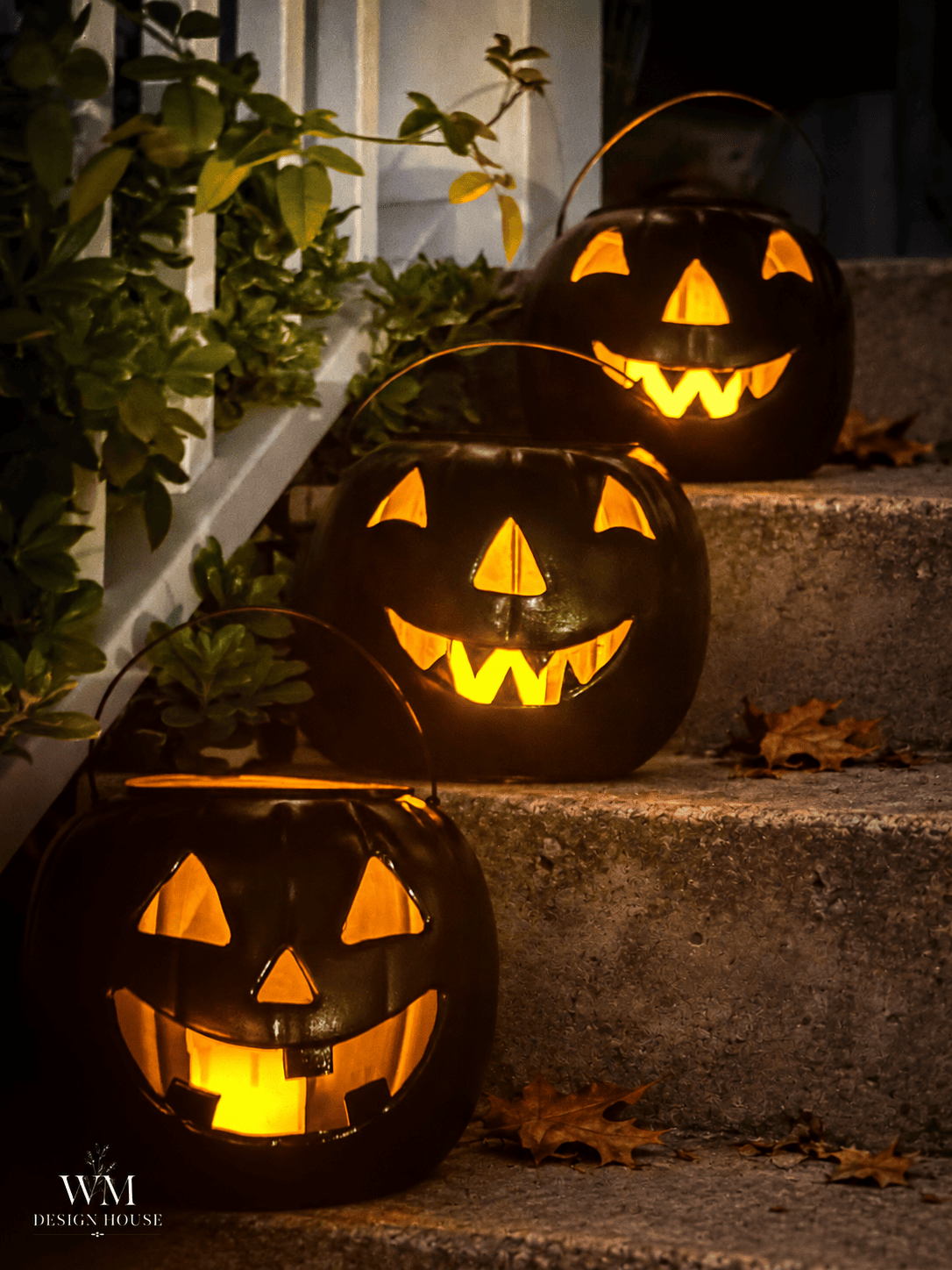 Three glowing halloween lanterns diy jack-o’-lanterns sit on concrete steps beside autumn leaves and green plants, creating a festive Halloween atmosphere in the evening.