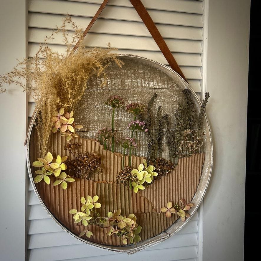 A circular wall hanging with dried grasses and flowers arranged on textured fabric, creating a natural, earthy scene. The piece hangs by a brown strap on white louvered closet doors.
