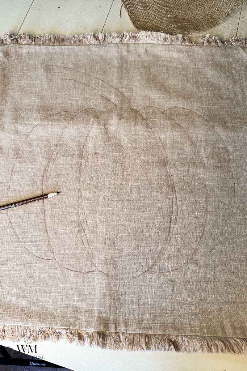 A pencil outline of a pumpkin is drawn on a rectangular piece of beige burlap fabric with frayed edges. A pencil rests on the left side of the fabric.