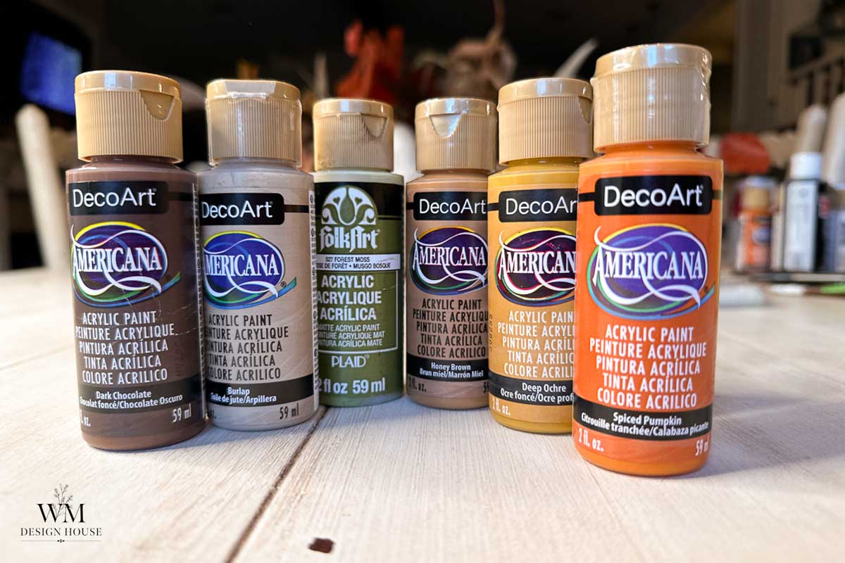 Six bottles of acrylic paint in various earth tones are lined up on a table. The brands are DecoArt Americana and FolkArt. Background is softly blurred, focusing on the paint bottles in the foreground.