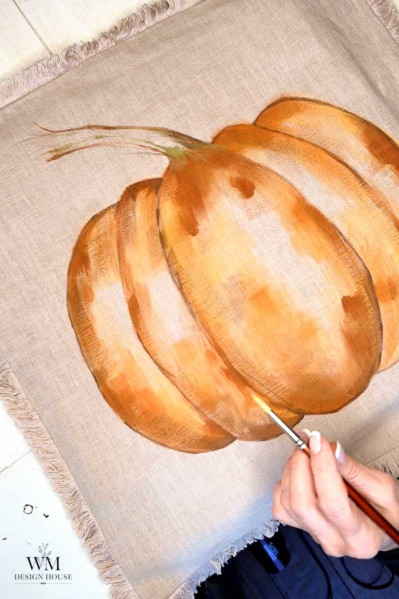 A hand is painting an orange pumpkin on a beige fabric with fringed edges. The pumpkin is partially complete, with brushstrokes visible. The WM Design House logo appears in the bottom left corner.