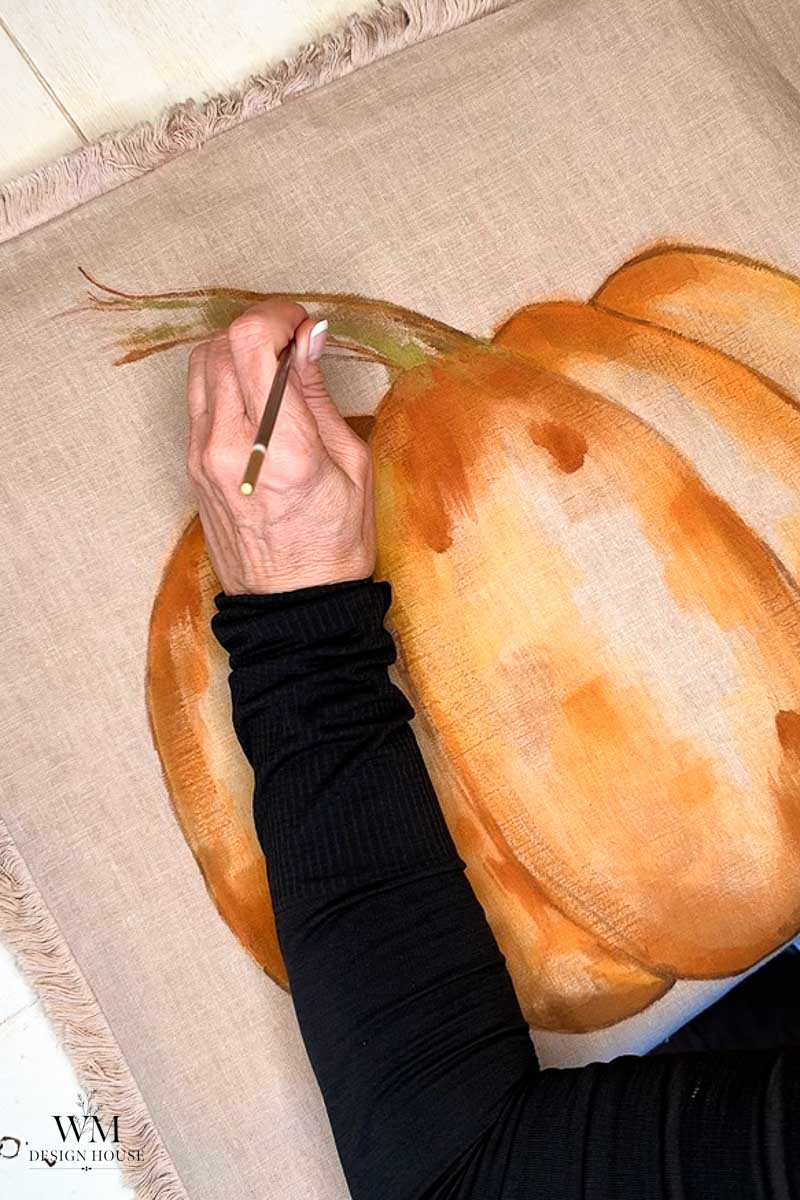 A person in a black sleeve paints the stem of a large orange pumpkin on beige fabric.