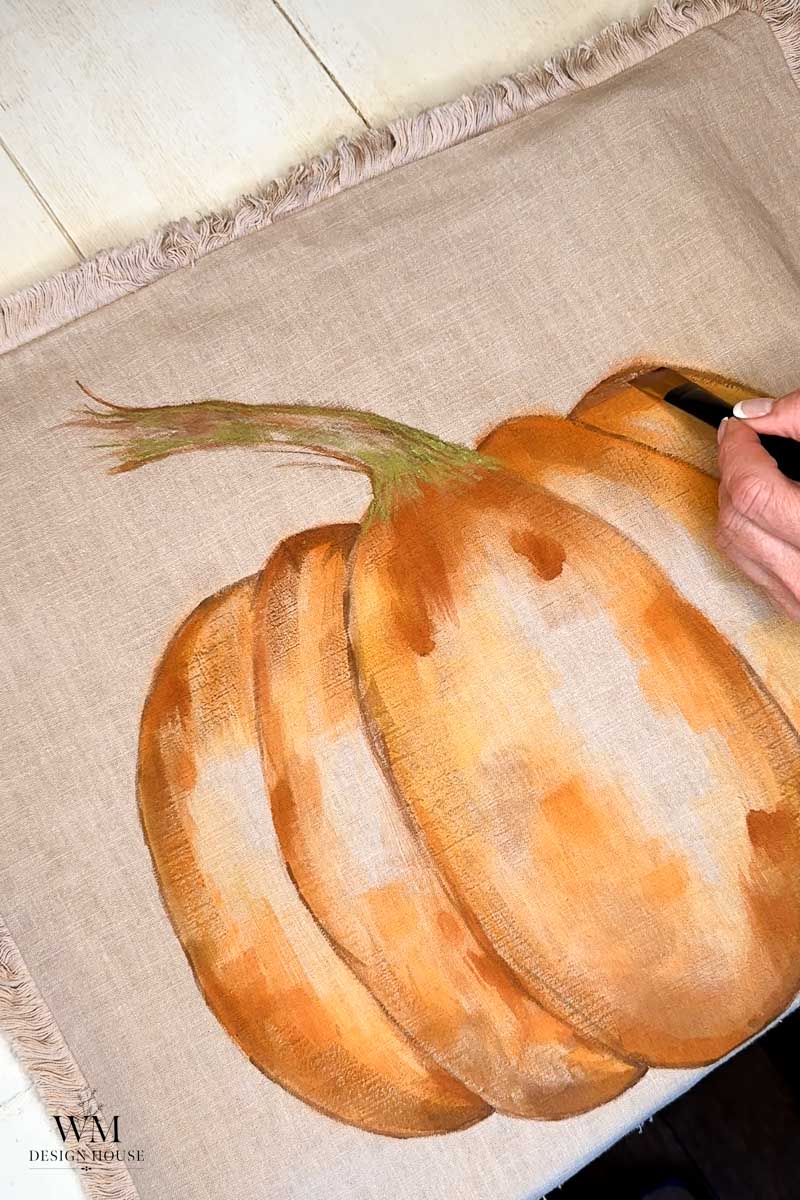 A hand paints an orange pumpkin on beige fabric with a black brush. The fabric has fringe edges and is laid on a light surface.