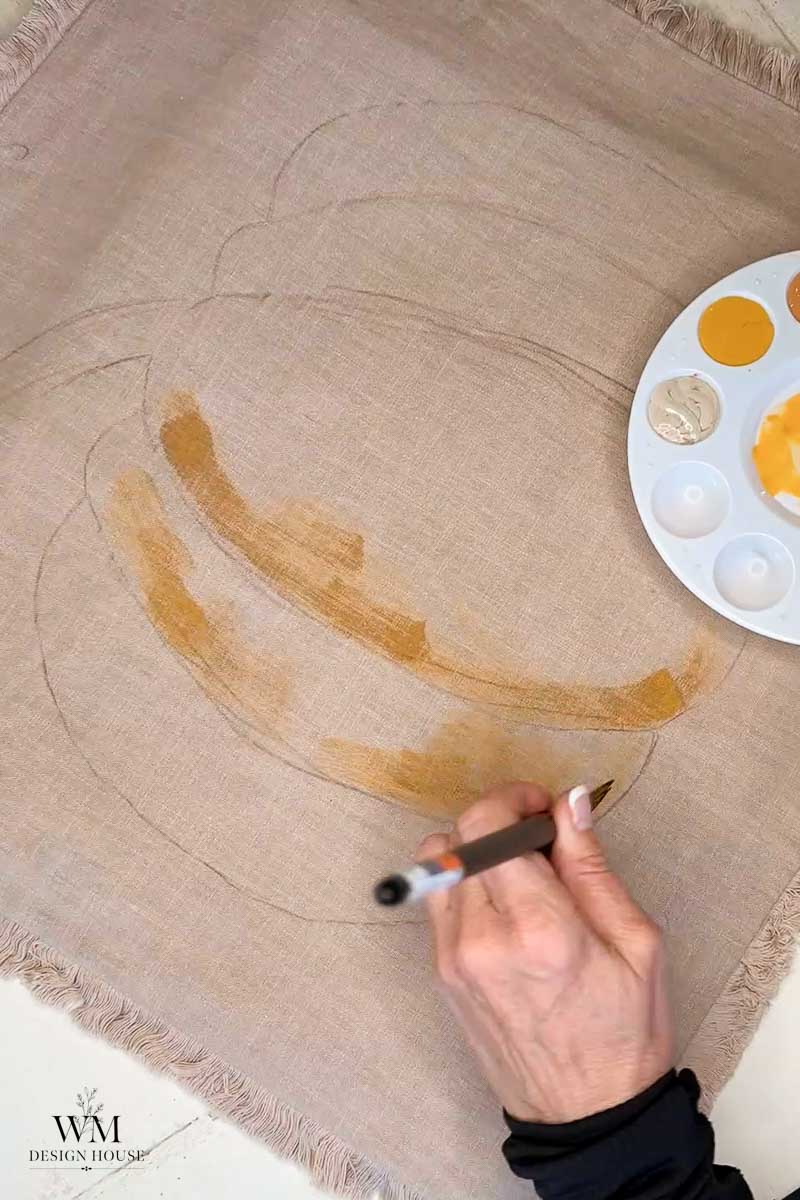 A hand begins painting a pumpkin outline on beige fabric using light brown and yellow paint from a palette.