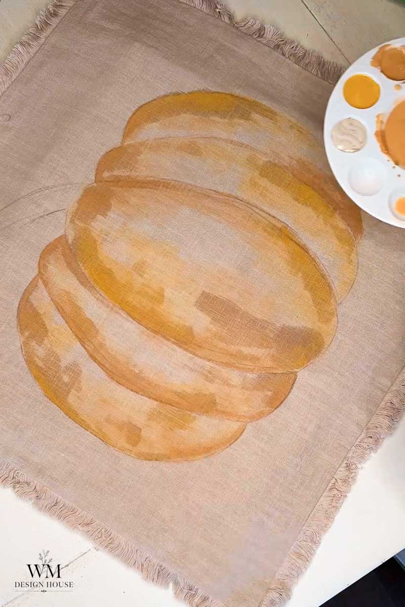 A painted fabric shows a stack of three golden-brown, oval-shaped bread slices. A palette with various paint shades sits at the top right corner. The fabric has fringed edges and WM Design House is marked in the bottom left.