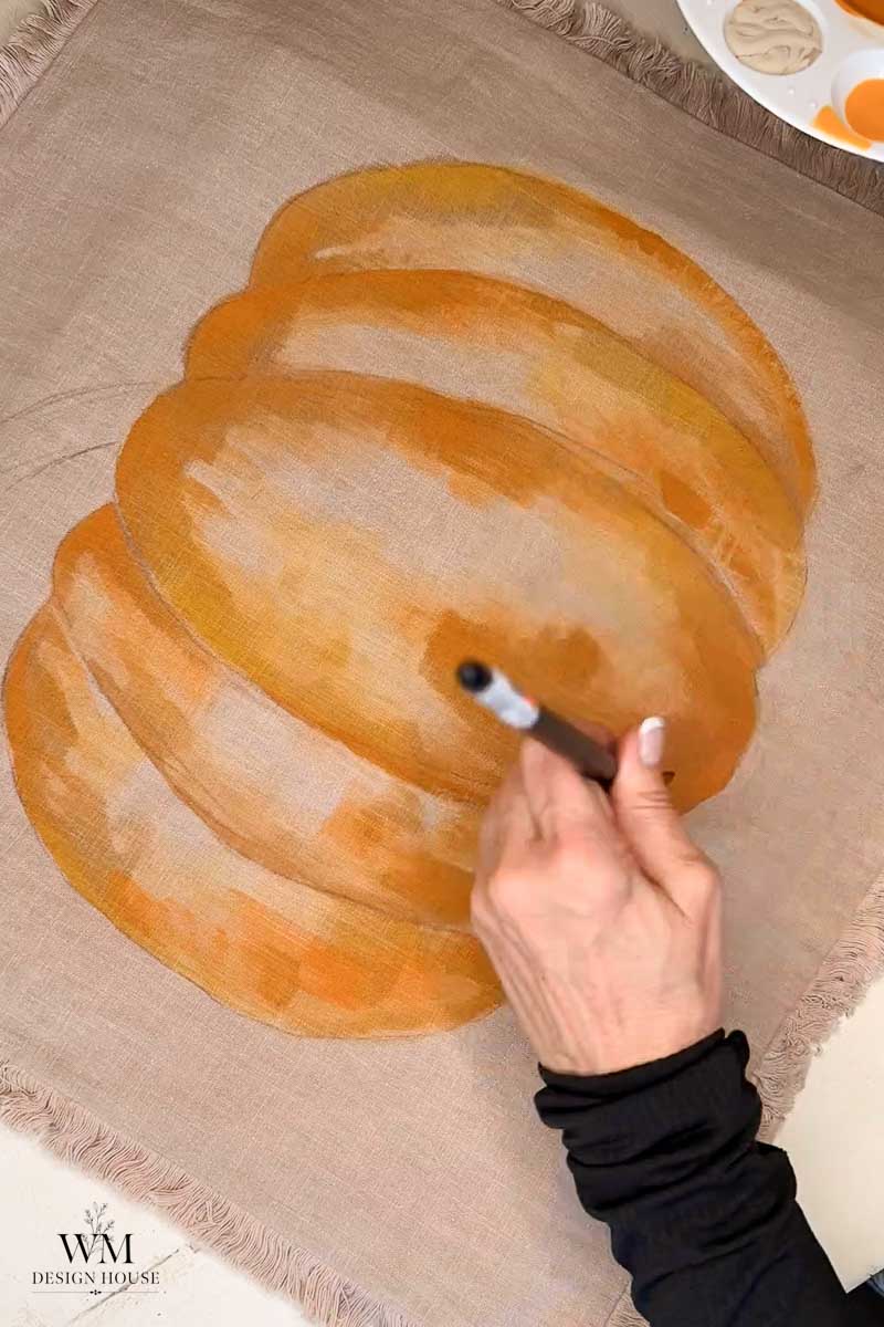 A person paints an orange pumpkin on beige fabric using a brush; a paint palette is visible in the corner.