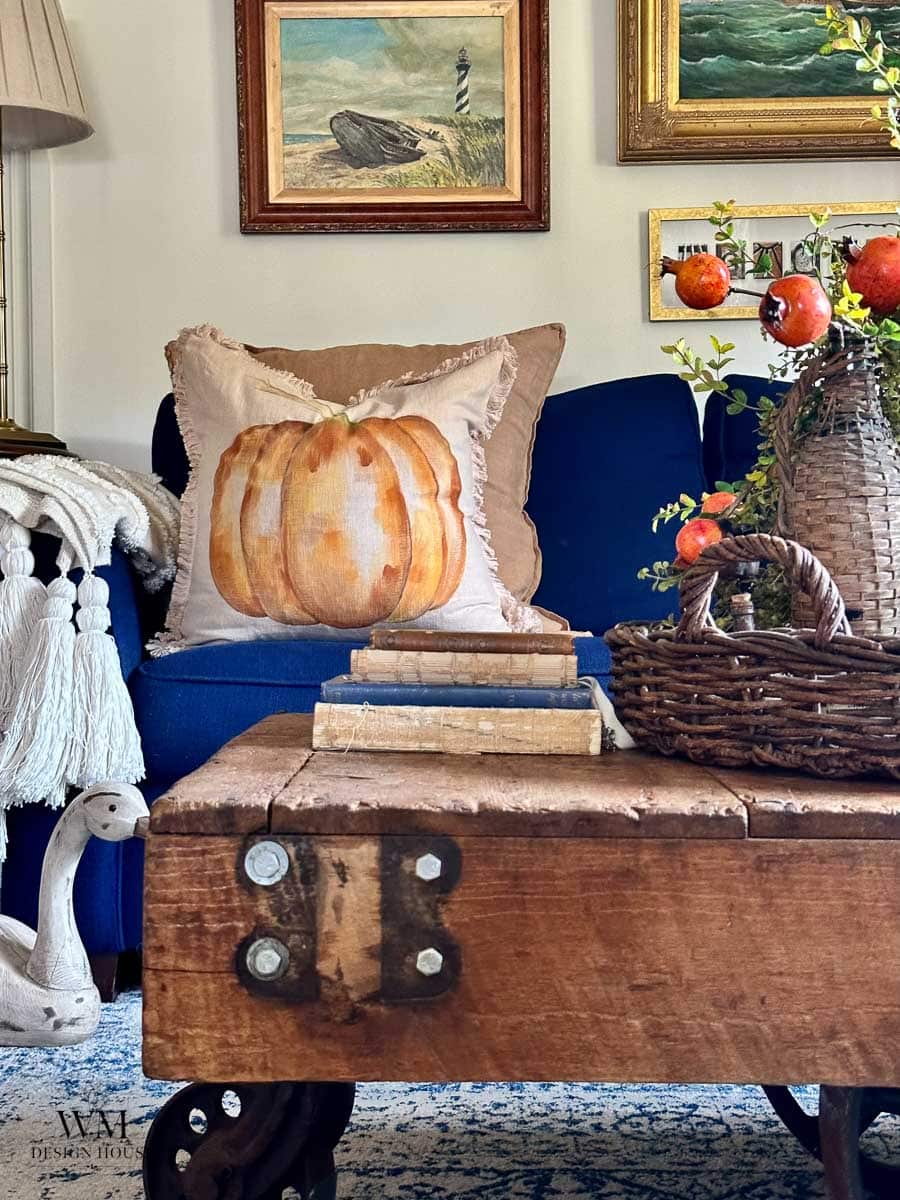 A cozy living room with a blue sofa, a pumpkin pillow, rustic wooden coffee table, stacked books, wicker basket with pomegranates, and framed artwork on the wall.
