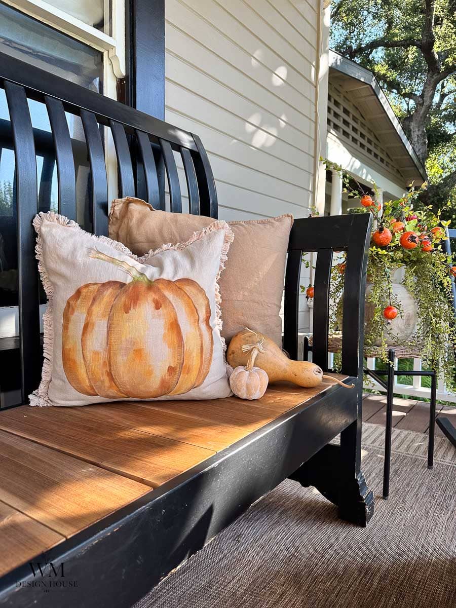 A black bench with autumn-themed pillows, including one with a pumpkin design, sits on a porch. Decorative gourds and a vibrant floral arrangement in a vase are nearby, creating a cozy fall atmosphere.