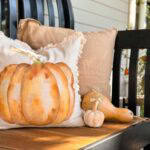 A black bench with tan pillows, one featuring a painted pumpkin. Two small gourds sit beside the pillows, and a basket with orange berries is nearby. The setting appears to be a porch decorated for autumn.