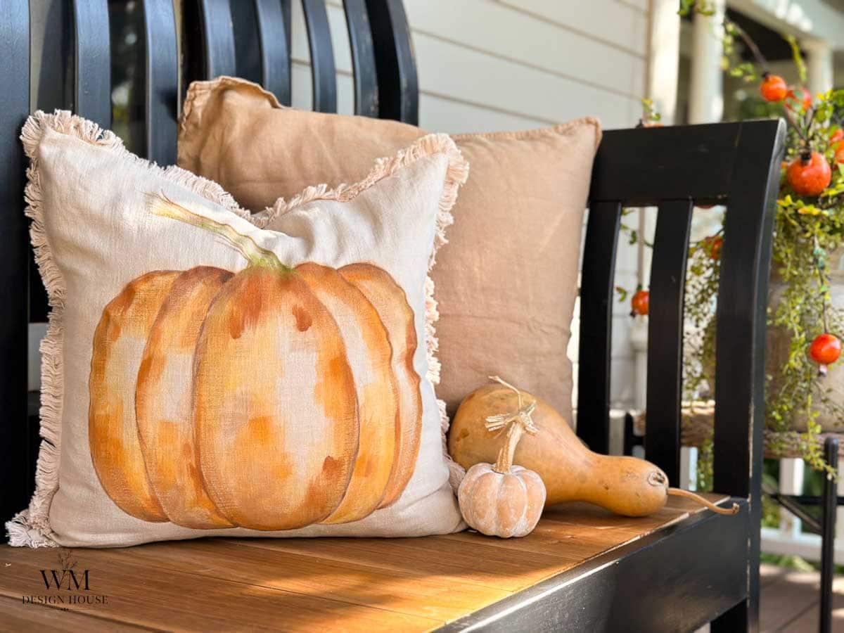 A black bench with tan pillows, one featuring a painted pumpkin. Two small gourds sit beside the pillows, and a basket with orange berries is nearby. The setting appears to be a porch decorated for autumn.