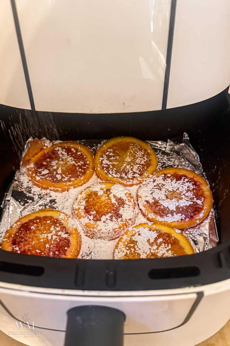 Six orange slices topped with powdered sugar are arranged on foil inside an air fryer. The orange slices appear slightly caramelized, and the air fryer lid is open, revealing the fruit inside.