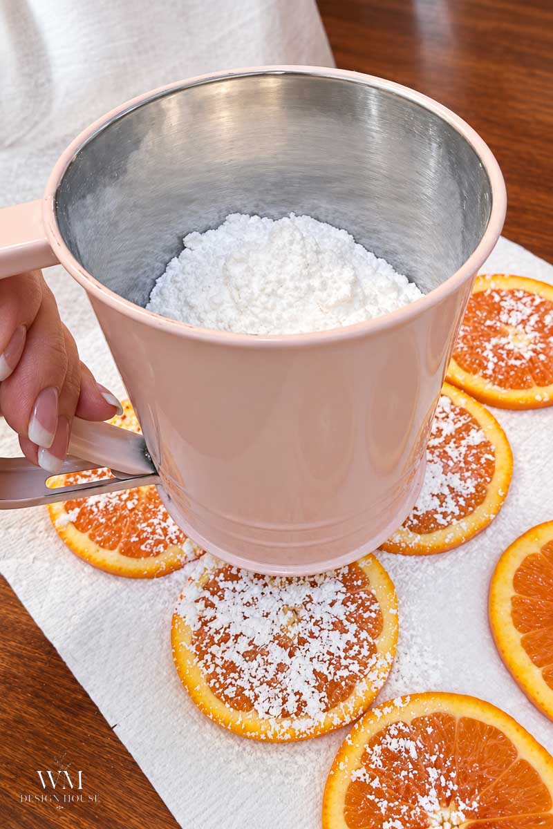 A hand holds a pink sifter with powdered sugar above orange slices on a paper towel, dusting them with sugar.