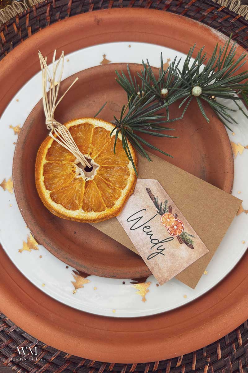 A holiday place setting with a terracotta plate, dried orange slice, pine sprig, and a brown name card reading “Wendy,” all on a white plate with gold stars and a woven charger underneath.