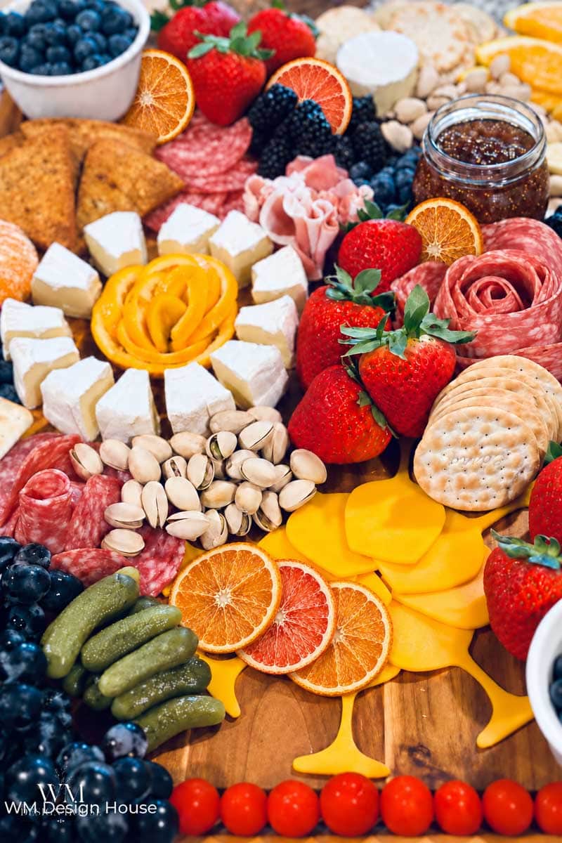 Charcuterie board with cheeses, crackers, strawberries, sliced citrus, meats, nuts, blueberries, blackberries, pickles, grape tomatoes, and a jar of jam on a wooden surface.