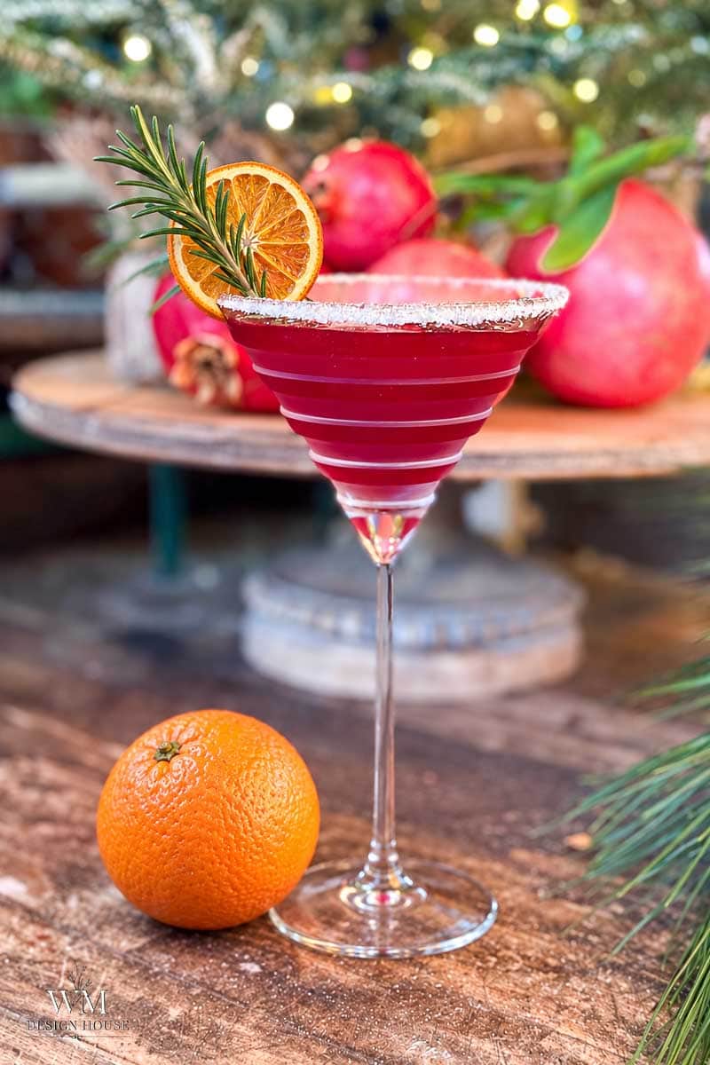 A bright red cocktail in a martini glass, garnished with a dried orange slice and rosemary sprig, sits on a wooden surface with a fresh orange and pomegranates in the background.