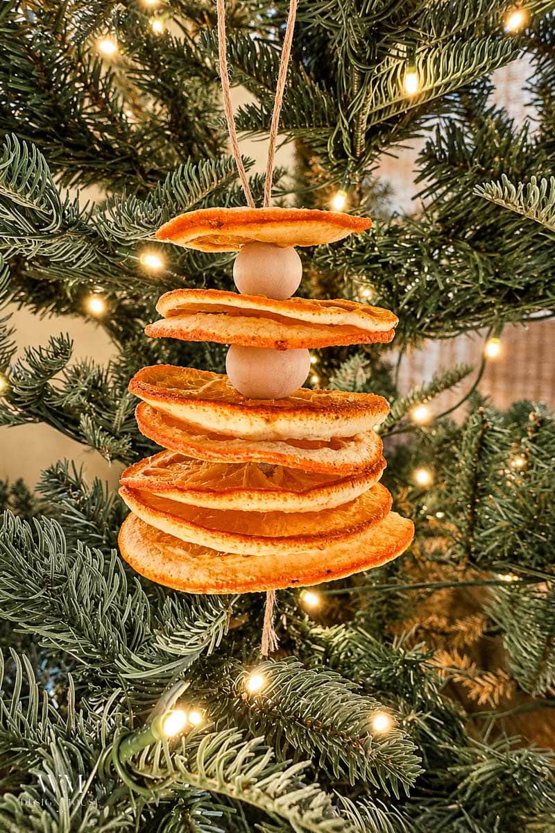 A Christmas tree decorated with an ornament made of dried orange slices and wooden beads, strung together and hung with twine, with warm string lights in the background.