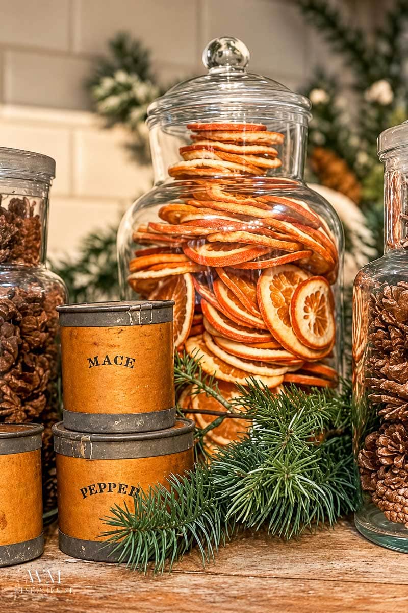 Glass jars filled with dried orange slices and cloves sit on a wooden surface, surrounded by small vintage spice tins labeled “MACE” and “PEPPER,” with pine branches adding a festive touch.