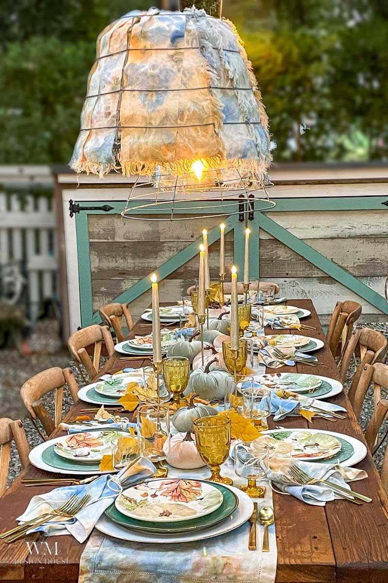 A rustic outdoor table is set for dinner with candles, yellow glassware, painted plates, pumpkins, and autumn decorations, capturing the essence of an elegant tablescape. A fringed fabric lampshade hangs above, casting a warm glow over this inviting fall tablescape.