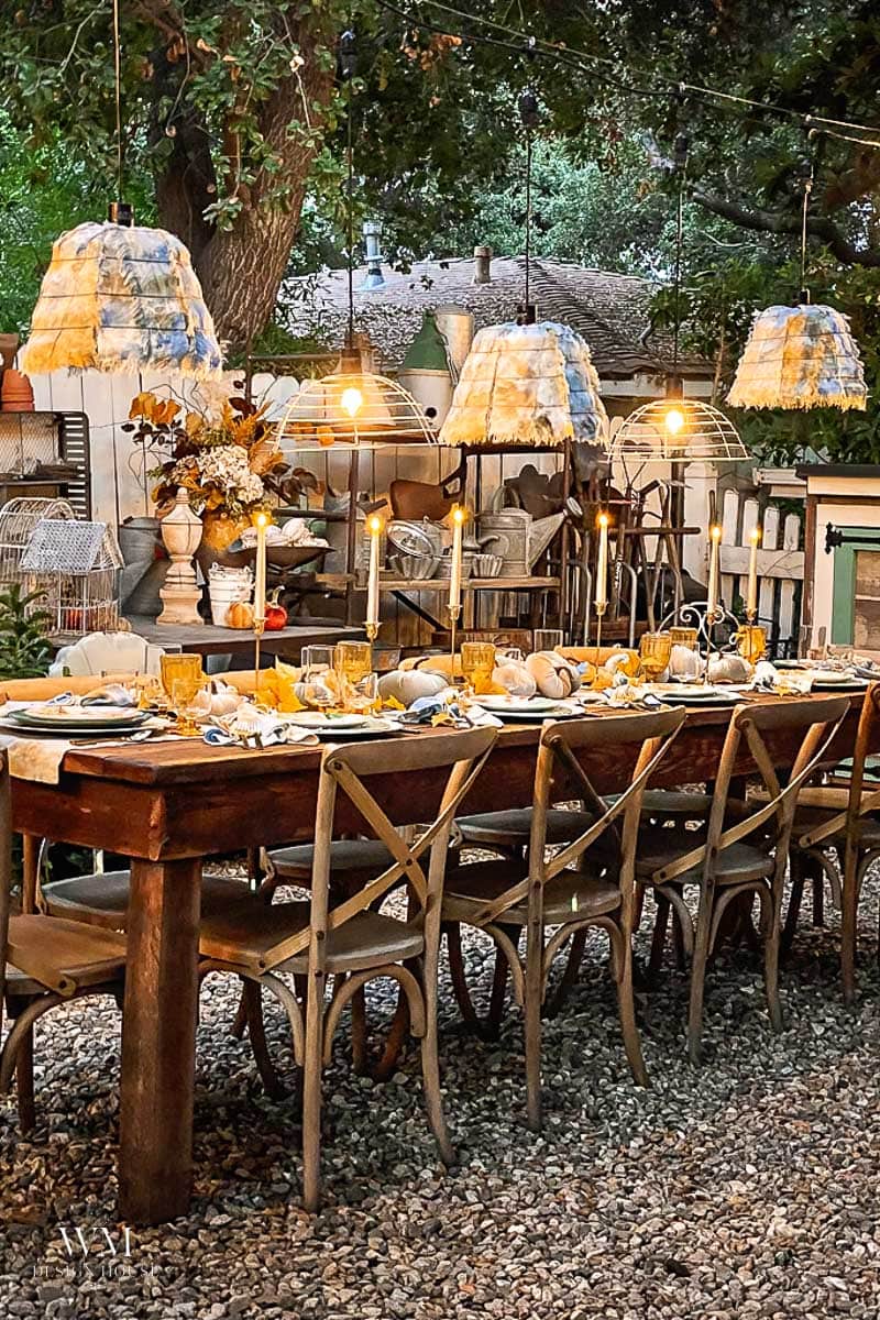 A long wooden outdoor dining table with ten chairs is set with candles, yellow glasses, and floral centerpieces, creating an elegant tablescape that is beautifully lit by hanging lamps and surrounded by trees and garden decor.