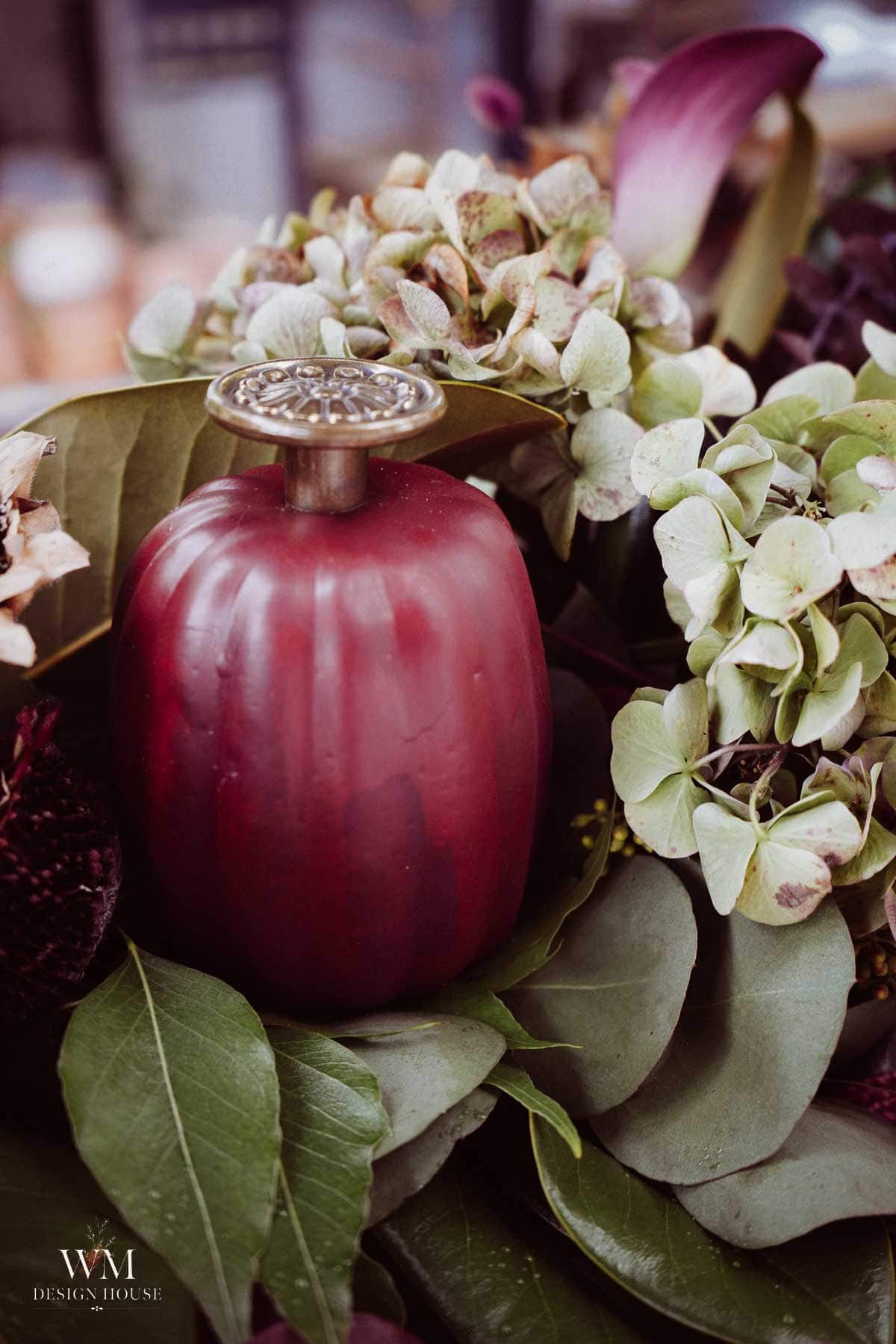 A red, apple-shaped decorative object with a metal top sits among green leaves and clusters of hydrangea flowers.